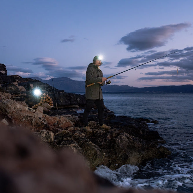A person stands on a shoreline at dusk fishing with a long rod, wearing a Ledlenser head torch that illuminates the area.