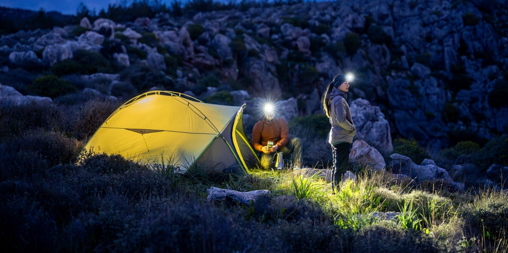 A camping tent set in a rocky outdoor landscape at night, with two people wearing Ledlenser head torches standing beside it in the foreground.