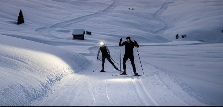 Two cross-country skiers wearing Ledlenser Head torches travel across a snowy mountain landscape.