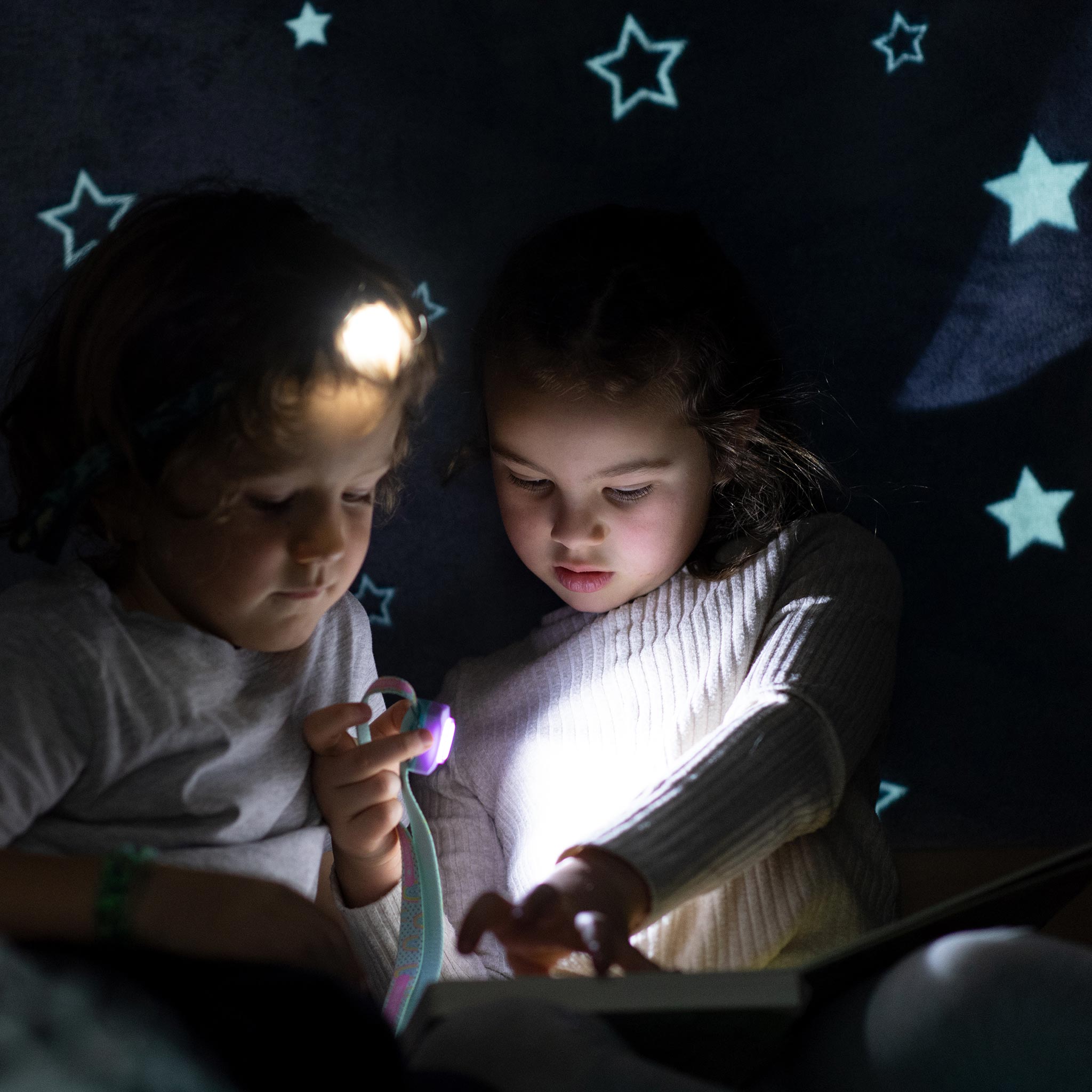Two children reading with a headtorch under a starry blanket